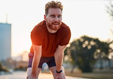 A smiling man with curly red hair and a beard leans forward, resting his hands on his knees. He wears a brown T-shirt and shorts, standing outdoors with a soft sunset light in the background. The scene conveys a sense of enjoyment and physical activity.