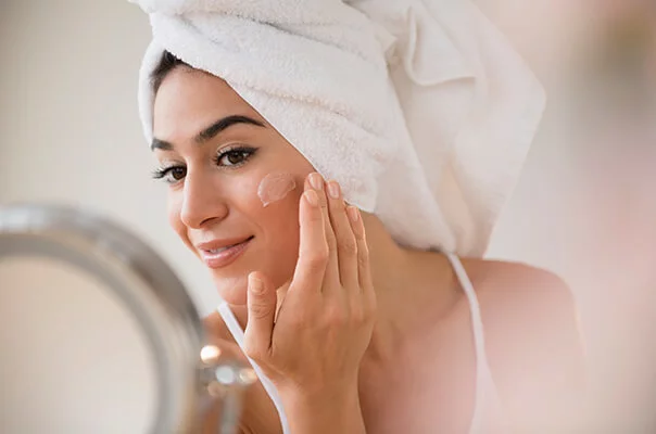 Frau mit weißem Handtuch auf dem Kopf steht vorm Spiegel und cremt sich das Gesicht ein